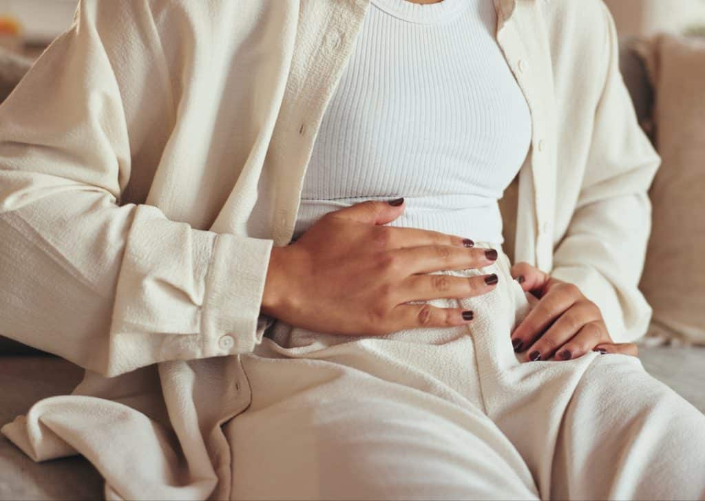 Mujer sujet&aacute;ndose el abdomen con dolor o molestias digestivas, vestida de color claro.
