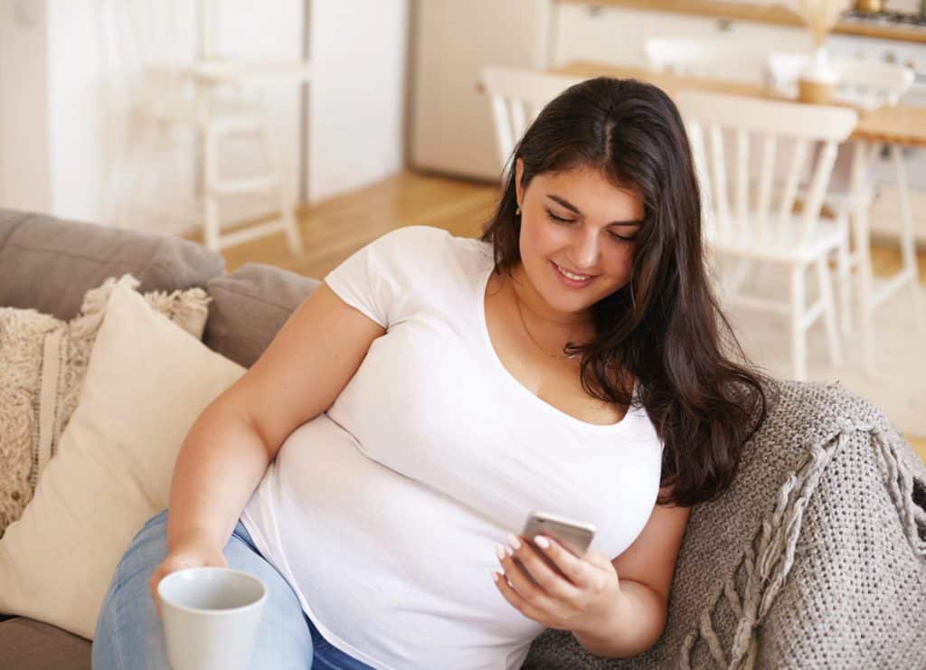 Mujer sentada en un sof&aacute; sonriendo mientras mira su tel&eacute;fono m&oacute;vil y sostiene una taza.