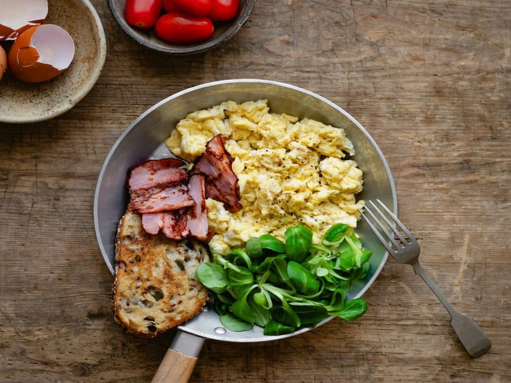 Sart&eacute;n r&uacute;stica con huevos revueltos, bacon y pan tostado sobre mesa de madera.