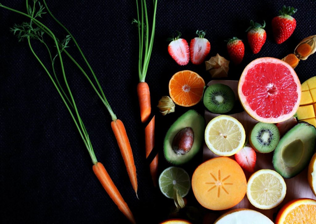 Bodegón de frutas y verduras ricas en vitaminas, incluyendo mango, aguacate, caqui, zanahorias y cítricos sobre fondo oscuro.