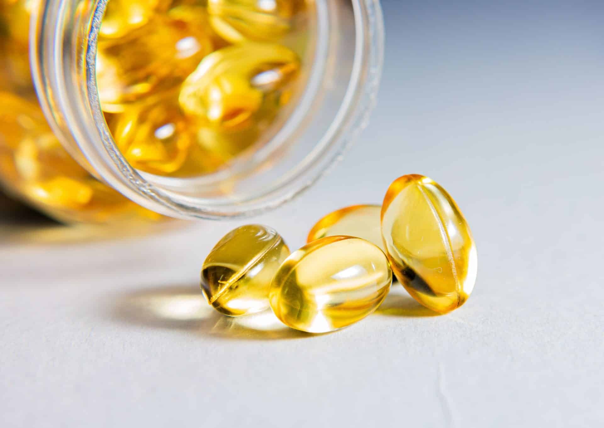 Cápsulas transparentes con líquido amarillo vertiéndose de un frasco de vidrio sobre fondo gris.