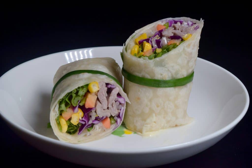 Dos mitades de un wrap integral relleno de tiras de pollo, col lombarda, ma&iacute;z y lechuga, servido en un plato hondo blanco sobre fondo oscuro.